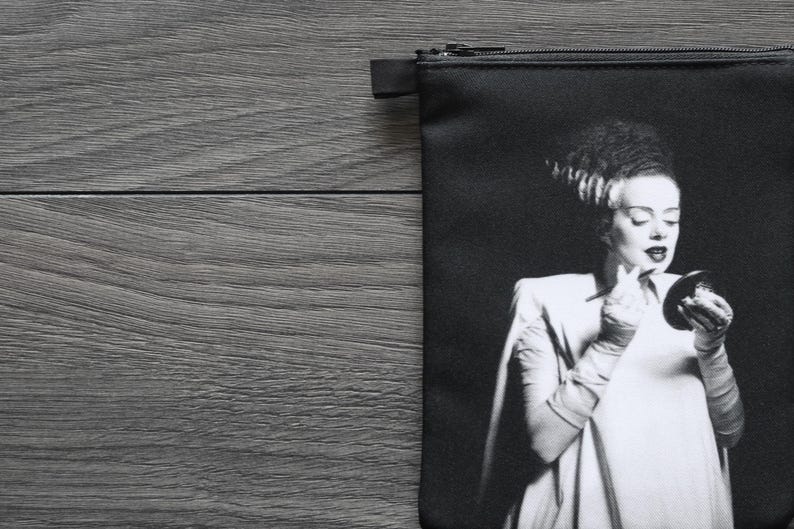 May include: Black zippered pouch featuring a black and white image of a woman in a white dress, holding a compact mirror. The image is a classic horror movie character. The pouch is on a gray wooden surface.