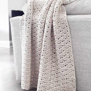 Puede incluir: Una manta de ganchillo beige sobre el brazo de un sofá gris. La manta tiene un patrón de tejido abierto y texturizado. El color neutro y la textura suave sugieren un artículo acogedor y hecho a mano para la decoración del hogar.