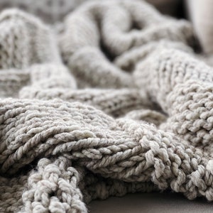 May include: Close-up of a soft, chunky knit throw blanket in a light beige colour. The blanket is made of thick yarn and has a textured, woven pattern.