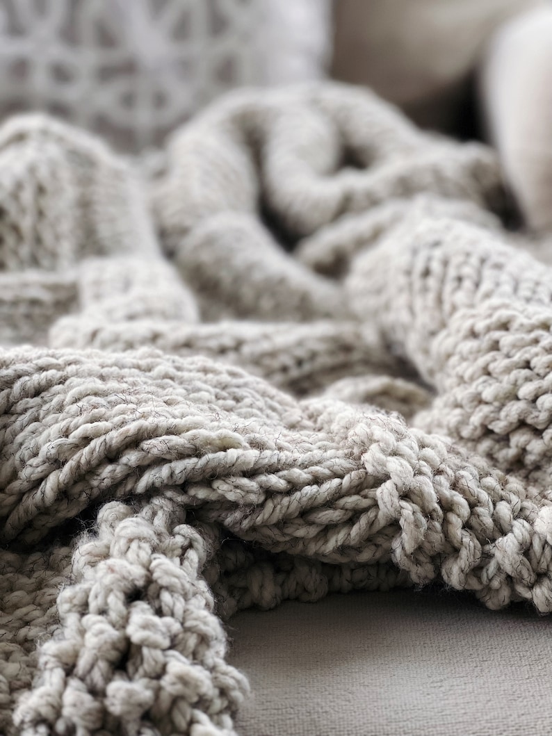 May include: Close-up of a soft, chunky knit throw blanket in a light beige colour. The blanket is made of thick yarn and has a textured, woven pattern.