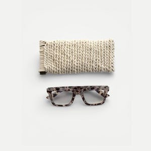 May include: A pair of eyeglasses with a speckled, light-colored frame and clear lenses, next to a cream-colored, knitted eyeglass case. The case is rectangular and has a woven texture.