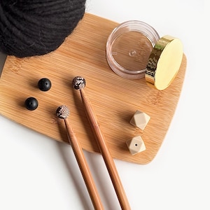 May include: A set of bamboo knitting needles with black and brown tips, a ball of black yarn, two small black beads, two beige silicone stitch markers, and a clear plastic container with a gold lid.