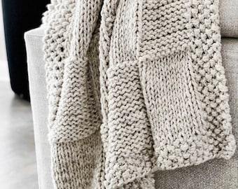 May include: A chunky knit throw blanket in a light beige color. The blanket is made with a textured stitch pattern and has a soft, cozy feel.