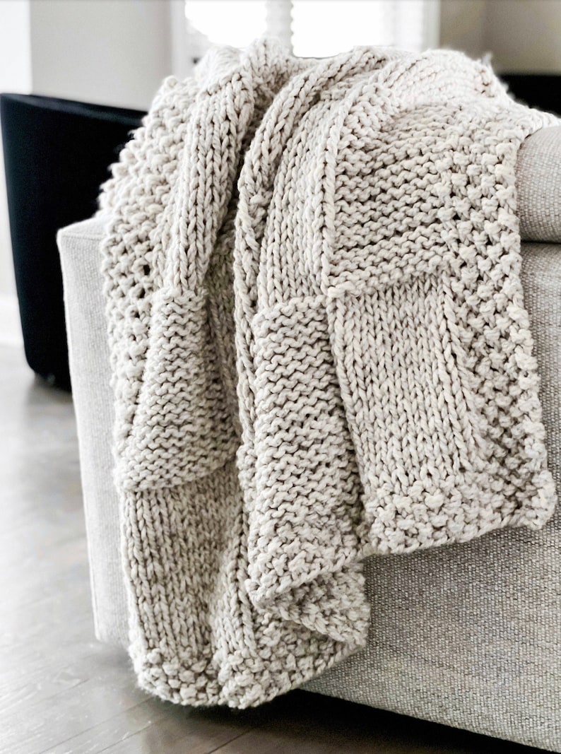 May include: A chunky knit throw blanket in a light beige colour. The blanket is made with a textured stitch pattern and has a soft, cosy feel.