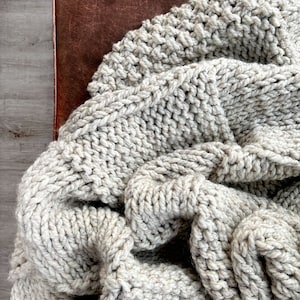 May include: A close-up of a chunky knit throw blanket in a light grey colour. The blanket is folded and draped over a brown leather surface.