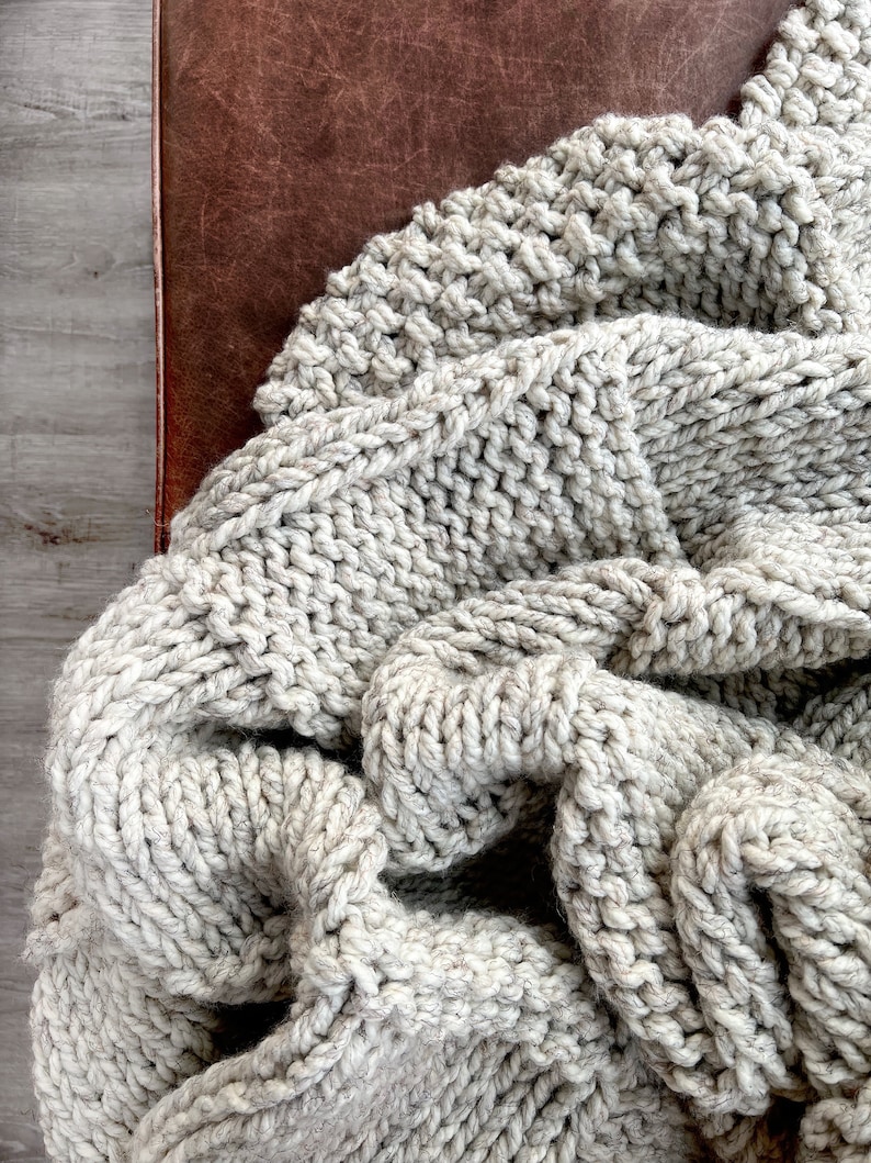 May include: A close-up of a chunky knit throw blanket in a light grey colour. The blanket is folded and draped over a brown leather surface.