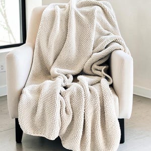 May include: A cream-colored knitted throw blanket draped over a beige armchair. The blanket has a textured, raised pattern and is folded to create soft, flowing lines. The chair has black legs.