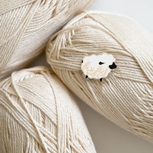Può includere: Uno spillo in smalto bianco a forma di pecora con occhi e zampe nere, circondato da foglie dorate. Lo spillo è sopra una palla di filato beige.
