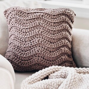 May include: A soft, mauve crocheted pillow with a wavy pattern sits on a white couch with a cream knitted throw blanket.