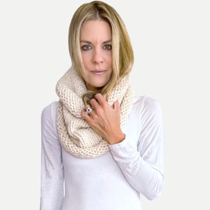 May include: A woman wearing a white turtleneck shirt and a cream-colored knitted cowl neck scarf. She is wearing blue jeans and has blonde hair.