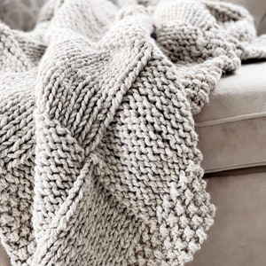 May include: A soft, chunky knit throw blanket in a light grey colour. The blanket is draped over a sofa and has a textured, cable knit pattern.