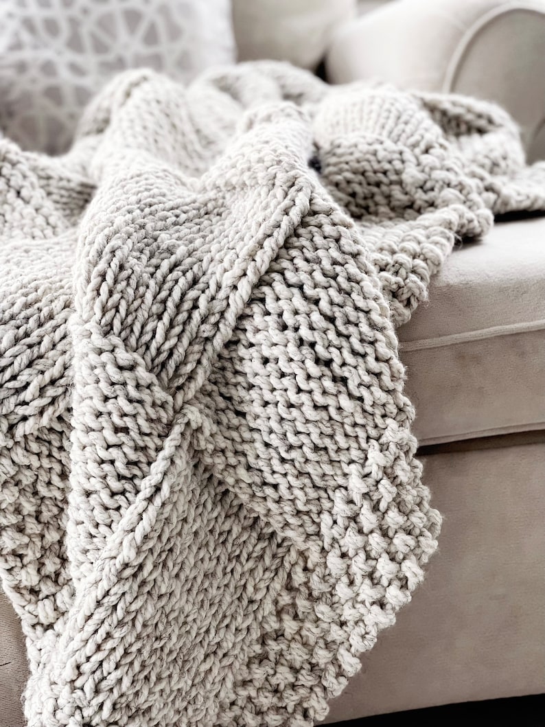 May include: A soft, chunky knit throw blanket in a light grey colour. The blanket is draped over a sofa and has a textured, cable knit pattern.