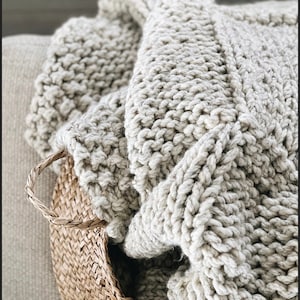 May include: A soft, chunky knit throw blanket in a light beige colour, folded and placed in a woven basket.