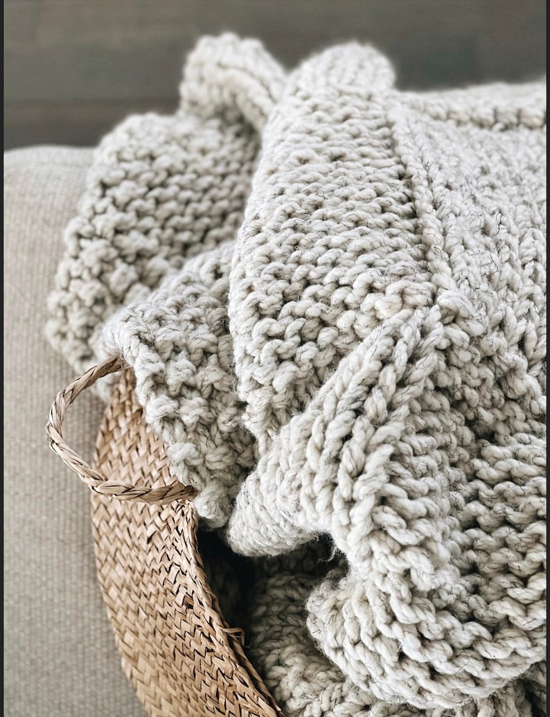May include: A soft, chunky knit throw blanket in a light beige colour, folded and placed in a woven basket.