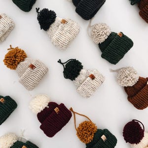 May include: A collection of miniature knitted beanies in various colors including dark green, cream, brown, burgundy, and black. Each beanie features a matching or contrasting yarn pom-pom on top, a folded brim, and a small brown rectangular tag.