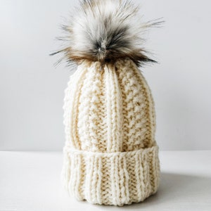 May include: Cream-colored knit beanie with a textured pattern and a ribbed brim. The hat is topped with a large, fluffy faux fur pom-pom in shades of brown, gray, and black. A cozy winter accessory.