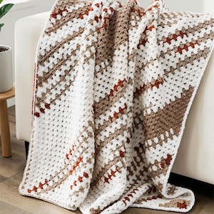 May include: A handmade crochet blanket draped over a white sofa. The blanket features a geometric pattern with white, brown, and rust-colored squares. The cozy throw is a blend of neutral tones, perfect for home decor.