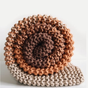 Op de afbeelding: Een rond gebreid kussen met drie concentrische cirkels van beige, oranje en bruin garen. Het kussen is gemaakt met een dikke breisteek.