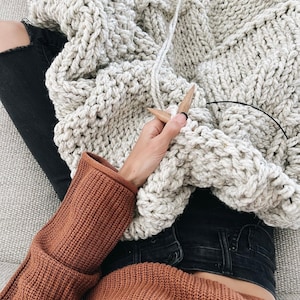 May include: A person knitting a large, chunky, cream-coloured blanket with wooden knitting needles. The person is wearing a rust-coloured sweater and black jeans.