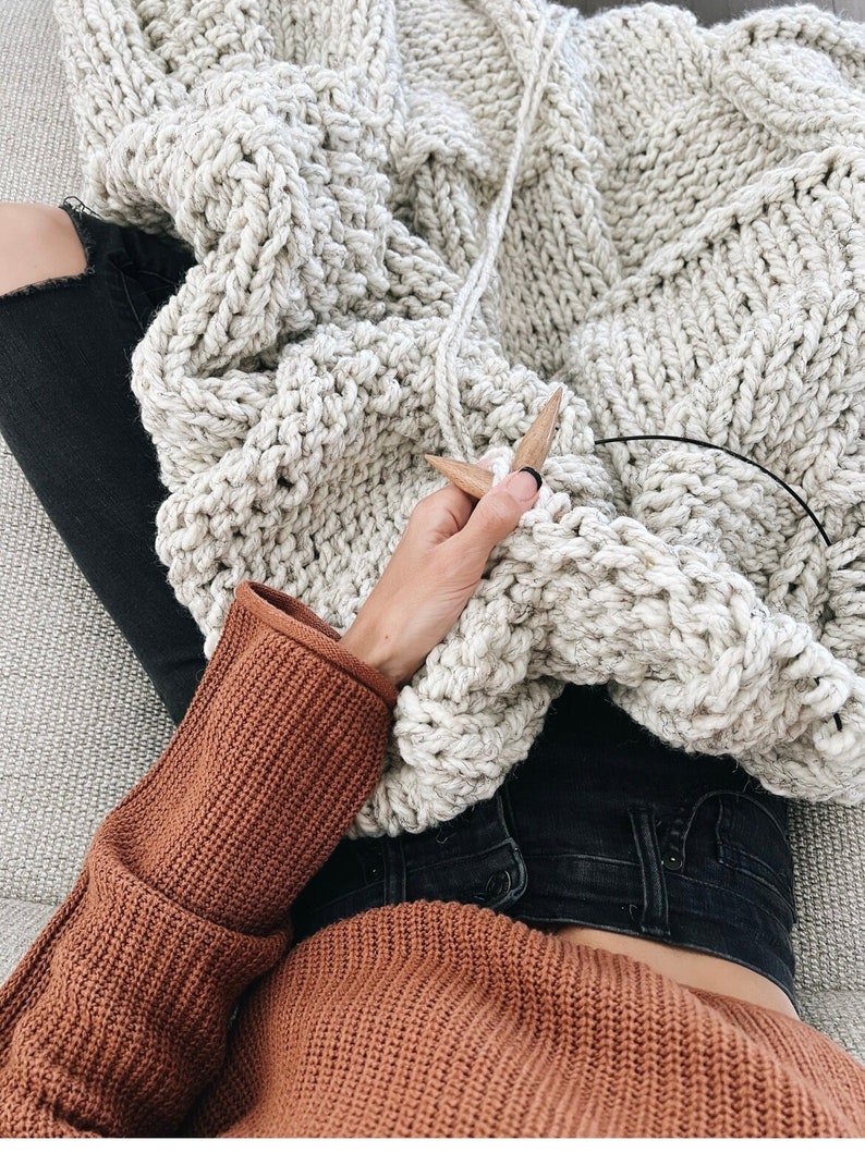 May include: A person knitting a large, chunky, cream-coloured blanket with wooden knitting needles. The person is wearing a rust-coloured sweater and black jeans.