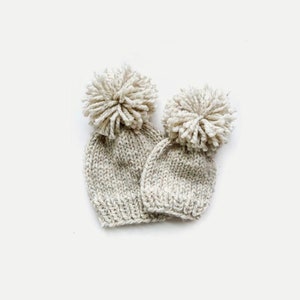 May include: Two knitted hats with pom-poms. The hats are beige with a subtle fleck pattern. The pom-poms are white.