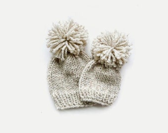 May include: Two knitted hats with pom-poms. The hats are beige with a subtle fleck pattern. The pom-poms are white.