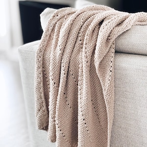 Könnte beinhalten: Eine weiche, beige gestrickte Wolldecke, die über ein Sofa gelegt ist. Die Decke hat ein zartes Lochmuster.