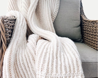 May include: A white crocheted throw blanket draped over a wicker chair with a gray cushion. The blanket has a ribbed texture and is made with a soft yarn.
