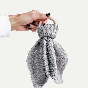 May include: A gray knitted baby teether with a wooden ring. The teether is soft and textured, making it perfect for babies to explore and chew on.