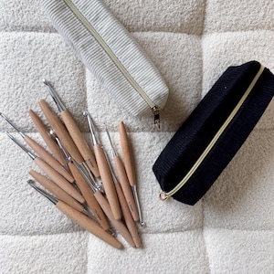 Puede incluir: Una colección de ganchillos de metal con mangos de madera clara, algunos marcados con tamaños como 10.0mm y 3.5mm, junto a dos estuches rectangulares con cremallera de tela de pana, uno en crema y otro en negro, con cremalleras doradas.