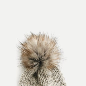May include: A beige knitted beanie with a fluffy brown pom-pom on top.