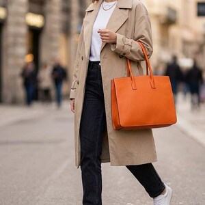 May include: A beige trench coat worn over a white t-shirt and black jeans, paired with white sneakers and a large, structured orange tote bag with two top handles. The outfit includes a simple gold chain necklace and small hoop earrings.