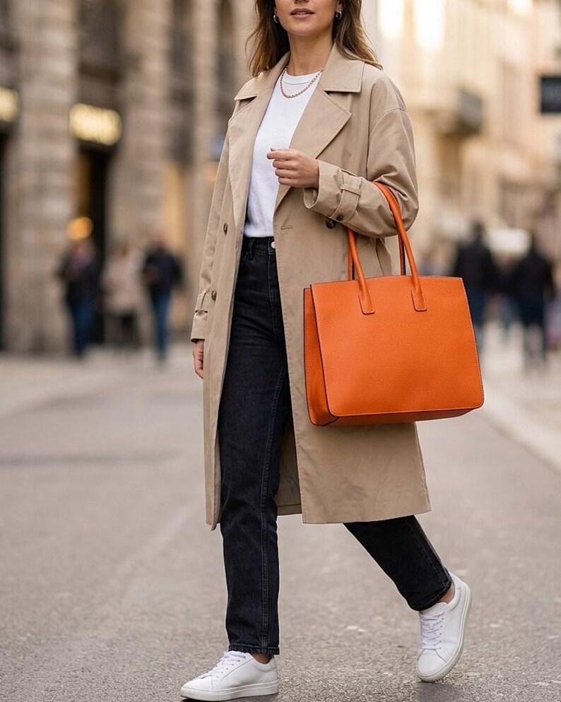 May include: A beige trench coat worn over a white t-shirt and black jeans, paired with white sneakers and a large, structured orange tote bag with two top handles. The outfit includes a simple gold chain necklace and small hoop earrings.