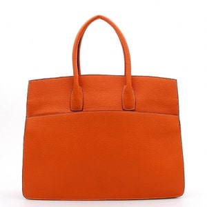 May include: A bright orange handbag with a textured surface, featuring two top handles and a structured rectangular body. The bag has visible stitching along the edges and a clean, minimalist design with a flat base and a wide opening at the top.