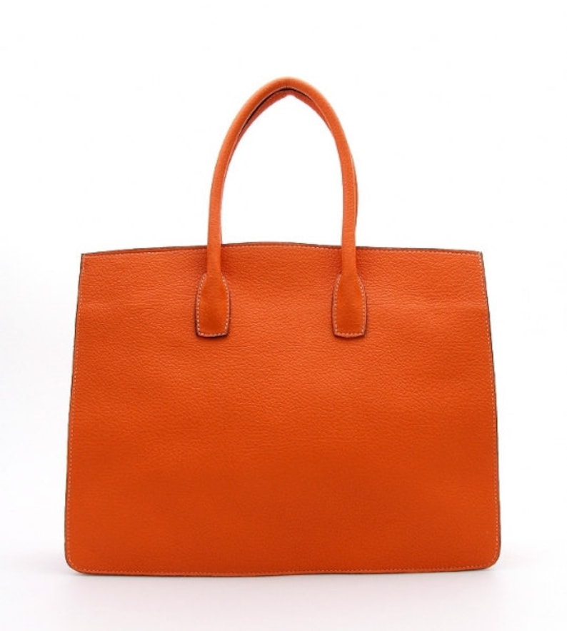 May include: A bright orange tote bag with a textured surface, featuring two matching top handles attached to the main body. The bag has a rectangular shape with clean, straight edges and visible stitching along the perimeter and handle bases.