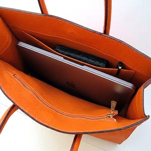 May include: An open orange textured leather tote bag with two top handles. The interior features a large main compartment containing a silver laptop with an Apple logo, a small black textured wallet, and a black pen. A zippered pocket is on the front.