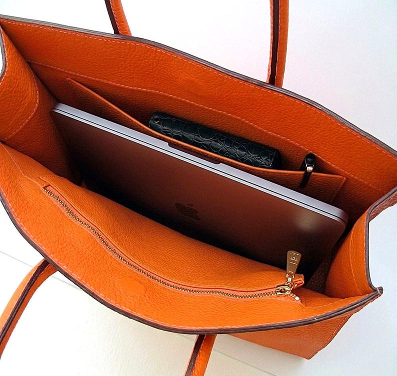 May include: An open orange textured leather tote bag with two top handles. The interior features a large main compartment containing a silver laptop with an Apple logo, a small black textured wallet, and a black pen. A zippered pocket is on the front.