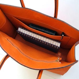 May include: An open orange tote bag with a textured leather-like surface. The interior features a large main compartment, a zippered pocket with a gold-toned pull, a slip pocket containing a dark green wallet, and a spiral-bound notebook.
