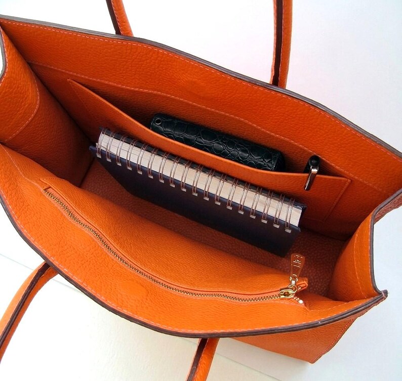 May include: An open orange tote bag with a textured leather-like surface. The interior features a large main compartment, a zippered pocket with a gold-toned pull, a slip pocket containing a dark green wallet, and a spiral-bound notebook.