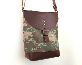 May include: A brown and camouflage crossbody bag with a flap closure. The bag features a brown leather flap, base, and strap, with a camouflage pattern on the main body. The bag has a buckle closure and is hanging against a white background.