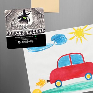 May include: A black square magnet featuring a drawing of a musical stage with a green witch and a crowd of people. The text "No One Mourns The Wicked" is on the magnet. A Spotify code is below the text.