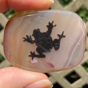 May include: A polished, oval-shaped stone with a translucent, banded appearance in shades of cream, tan, and white. A black silhouette of a frog is centered on the stone. The stone is being held up by a person.