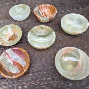 以下が含まれることがあります： さまざまな形とサイズの7つの小さな装飾用オニキスボウルのコレクション。ボウルは、緑、茶色、白、赤など、さまざまな色で、ユニークな模様と静脈があります。ボウルは暗い木製の表面に配置されています。