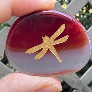 May include: A smooth, oval-shaped stone with a gradient of red to lavender hues. A gold-colored dragonfly design is centered on the stone. The stone is being held up by a person's fingers.