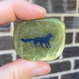 May include: A green, rectangular stone with a black fox silhouette. The stone is held between fingers and has a slightly rounded shape. The fox is depicted in a side profile, with a detailed outline.