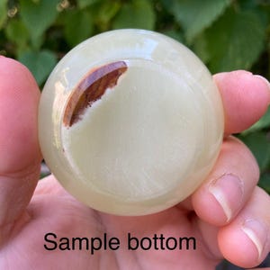 以下が含まれることがあります： 手で持たれた、茶色のインクルージョンが入った淡い緑色の球体。球体は滑らかな表面で、「Sample bottom」というテキストが画像の下部に表示されています。