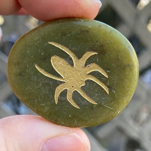 May include: A green, oval-shaped stone with a gold spider design. The spider is centered on the stone and has eight legs. The stone is held between fingers.
