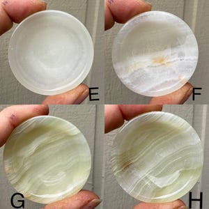 以下が含まれることがあります： 白と緑のさまざまな色合いの、丸く磨かれたオニキスストーンが4つ表示されています。各石には、渦巻きと帯のユニークなパターンがあり、一部には微妙な黄色または茶色のインクルージョンがあります。石は指で持たれています。