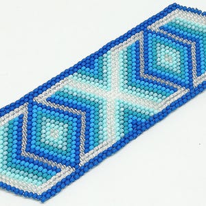 May include: A beaded bracelet featuring a geometric design in shades of blue, turquoise, and white. The bracelet is rectangular with a silver clasp and a detailed pattern of squares and diamonds.