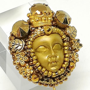 May include: A gold-toned brooch featuring a sculpted face with a crown and surrounding embellishments. The brooch is adorned with beads, floral accents, and leaf details. The design is set against a clear, rectangular base.